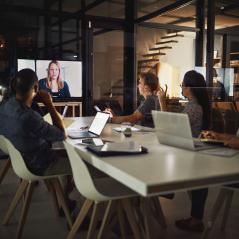 A group of people sitting at a table, watching a woman speaking on a screen at the end of the table